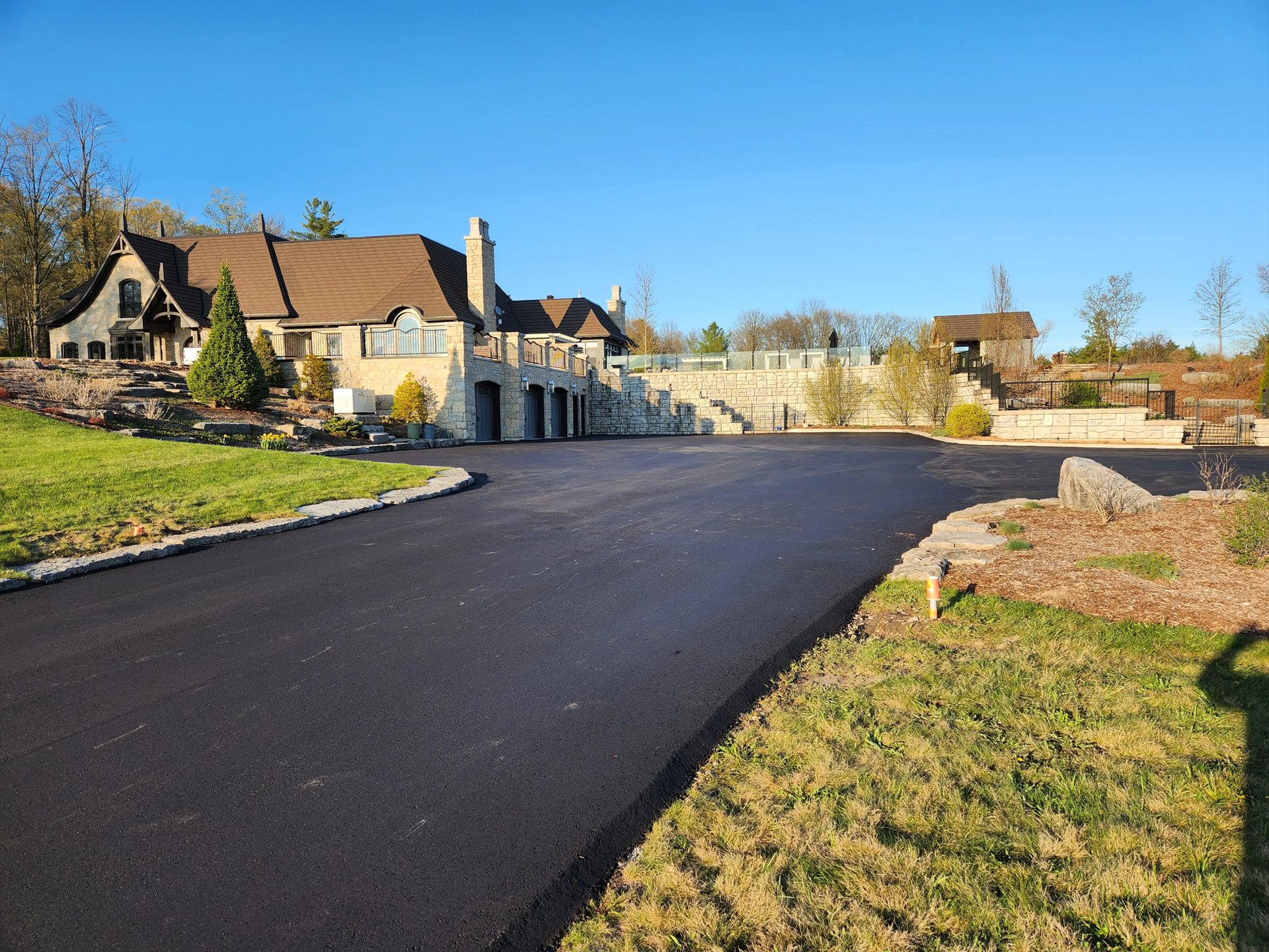 A broad finished asphalt driveway curves toward residential garages beside stone edging and landscaped beds.